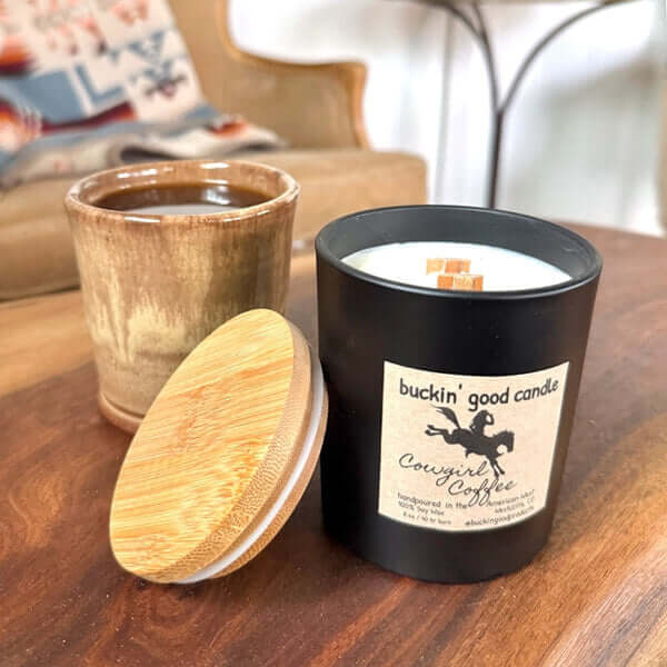 Candle labeled 'buckin' good candle Cowgirl Coffee' next to a wooden mug on a table.