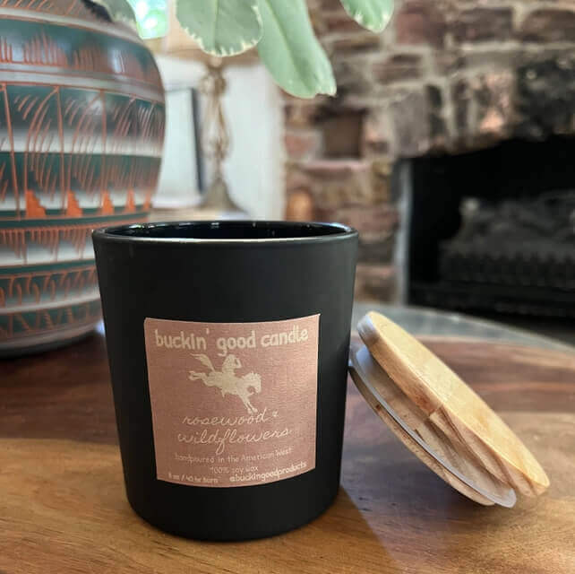 Black candle with a label on a wooden surface, with a vase and plant in the background.