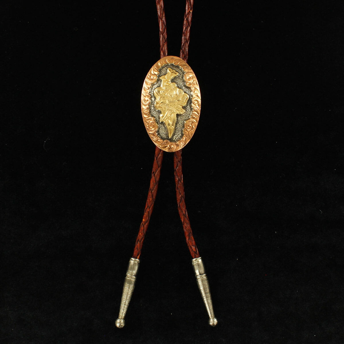 The oval bolo tie features an antiqued silver with copper floral trim and gold floral accent in the center with a brown leather cord. Measure 38” long with antiqued silver tips.