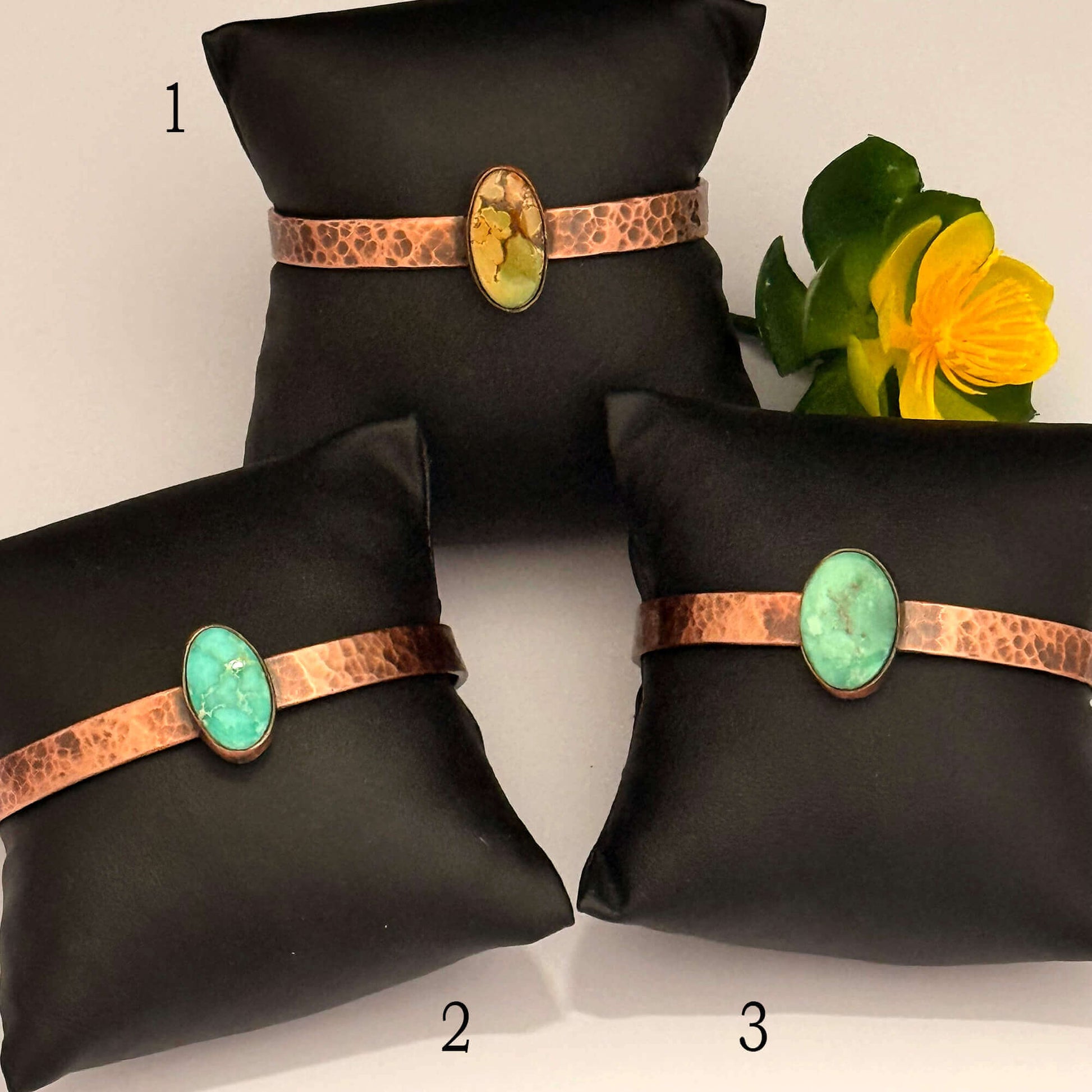 Three black jewelry display pillows with turquoise and copper accents, accompanied by a yellow flower.
