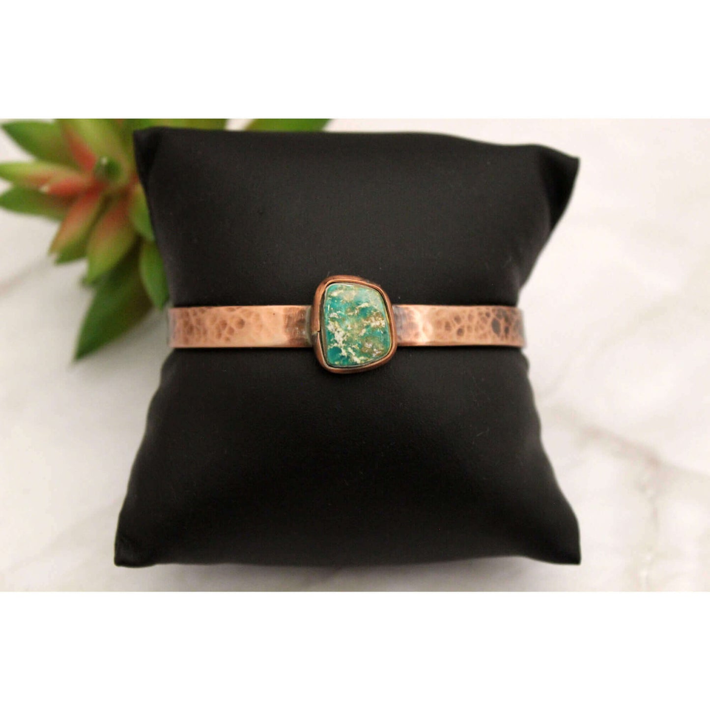 Bracelet with turquoise stone and copper band on a black pillow with a plant in the background