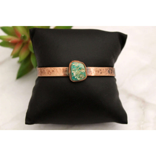 Bracelet with turquoise stone and copper band on a black pillow with a plant in the background