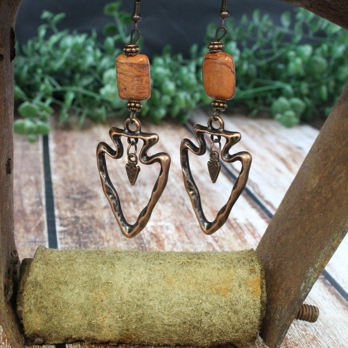 A pair of dangling earrings with rectangular orange-brown beads and hammered bronze arrowhead-shaped charms, displayed on a rustic wooden stand with blurred green foliage in the background.