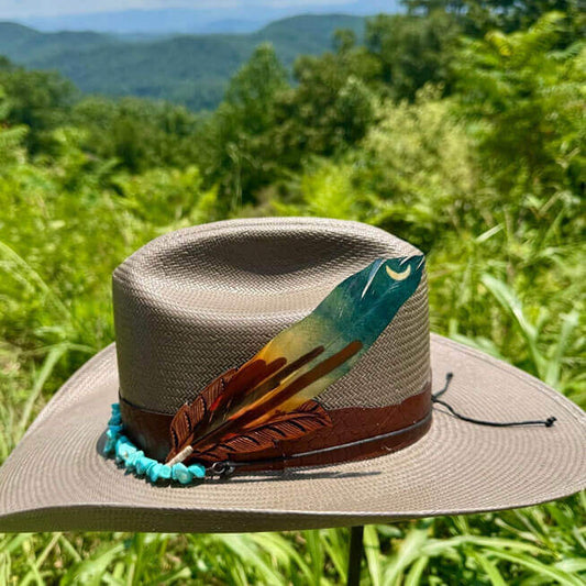 Beige straw hat with a decorative feather and beads against a green mountainous landscape.