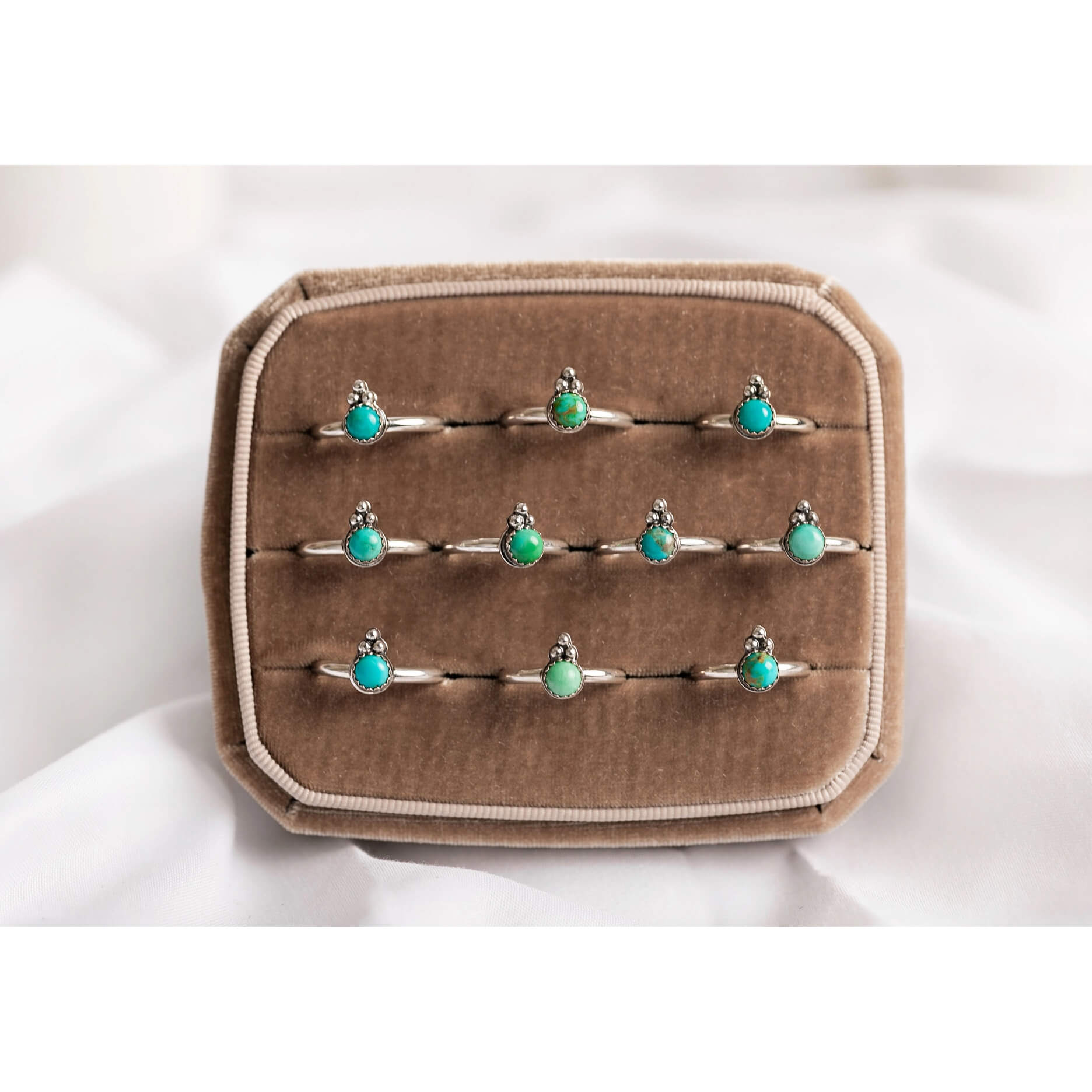 A set of nine silver rings with small oval turquoise-blue stones, displayed on a brown velvet ring box against a white fabric background.