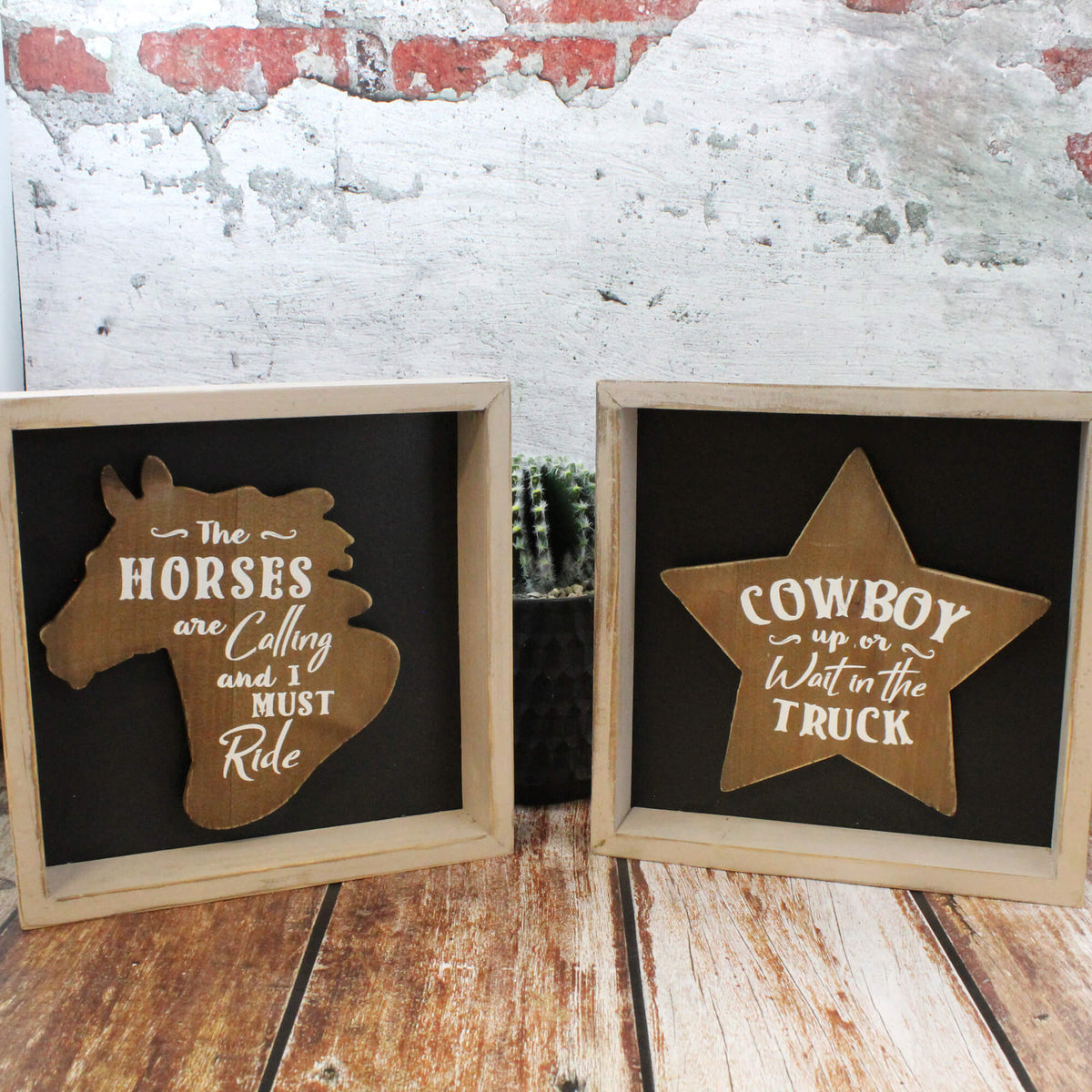 Two rustic framed cowboy-themed wall signs displayed side-by-side on a wooden surface against a distressed white and red brick wall. The left sign features a light wood horse silhouette with the text 'The Horses are Calling and I Must Ride' on a black background.
