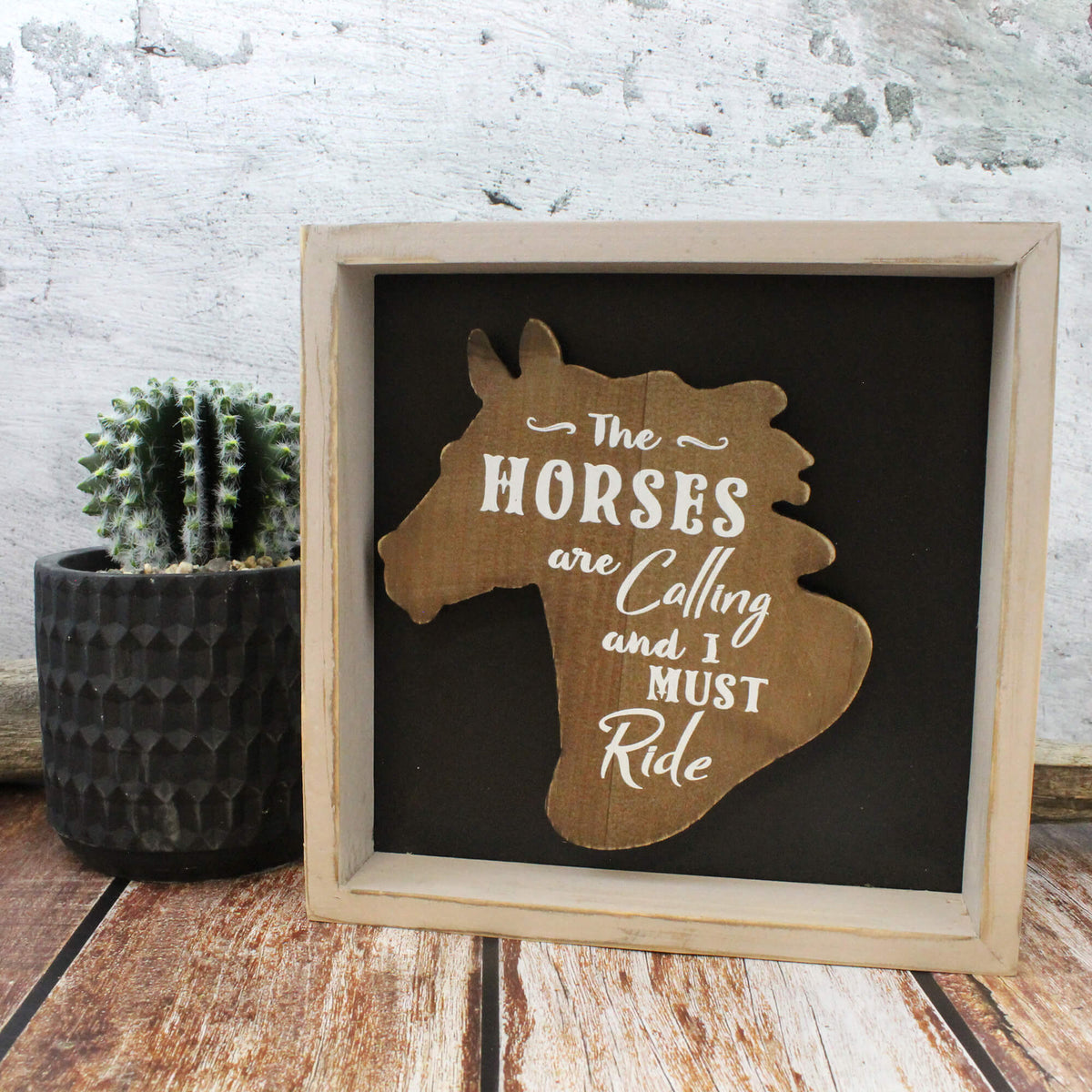 A square framed wooden sign with a horse silhouette and the phrase 'The HORSES are Colling and I MUST Ride' in white lettering on a dark background, styled on a rustic wood surface beside a small potted cactus.
