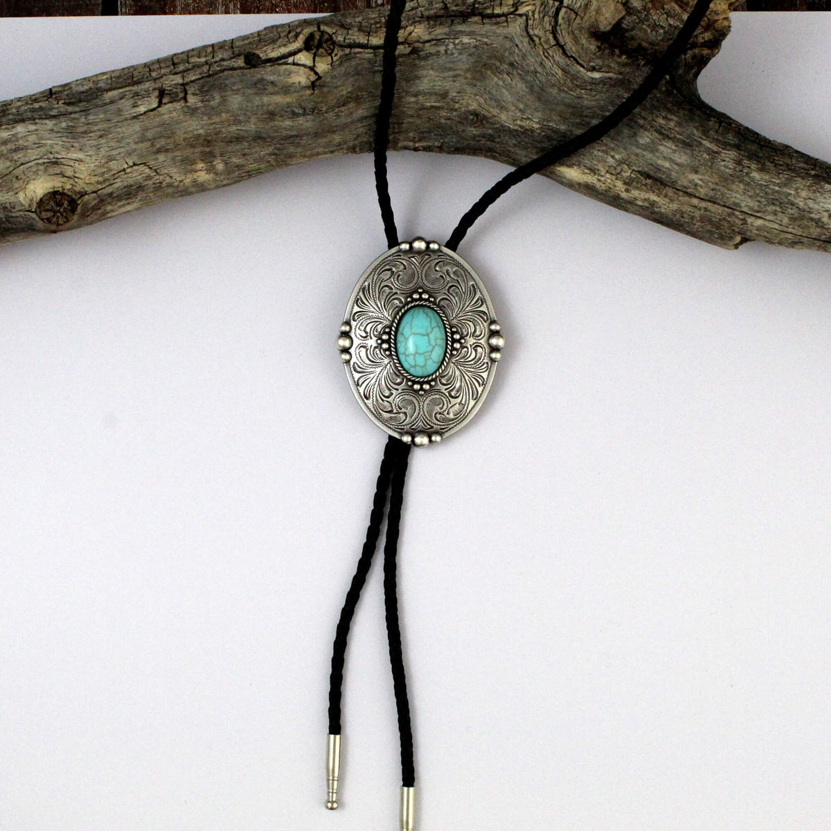 A silver-toned enlarged bolo tie necklace with an oval engraved pendant featuring a turquoise stone in the center, suspended on a black cord and displayed against a white background with a piece of driftwood.