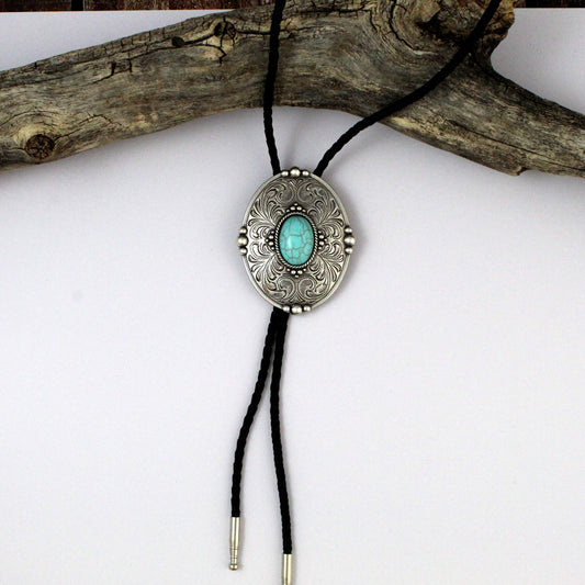 A silver-toned enlarged bolo tie necklace with an oval engraved pendant featuring a turquoise stone in the center, suspended on a black cord and displayed against a white background with a piece of driftwood.