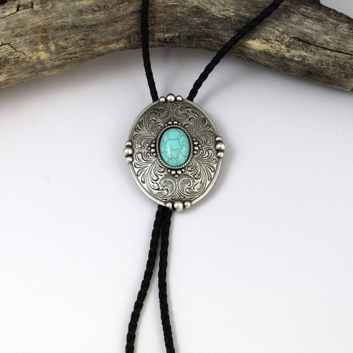 A silver-toned oval bolo-style pendant necklace with an engraved floral pattern, a turquoise-colored center stone, and black cords, displayed against a white background with a piece of weathered wood at the top.