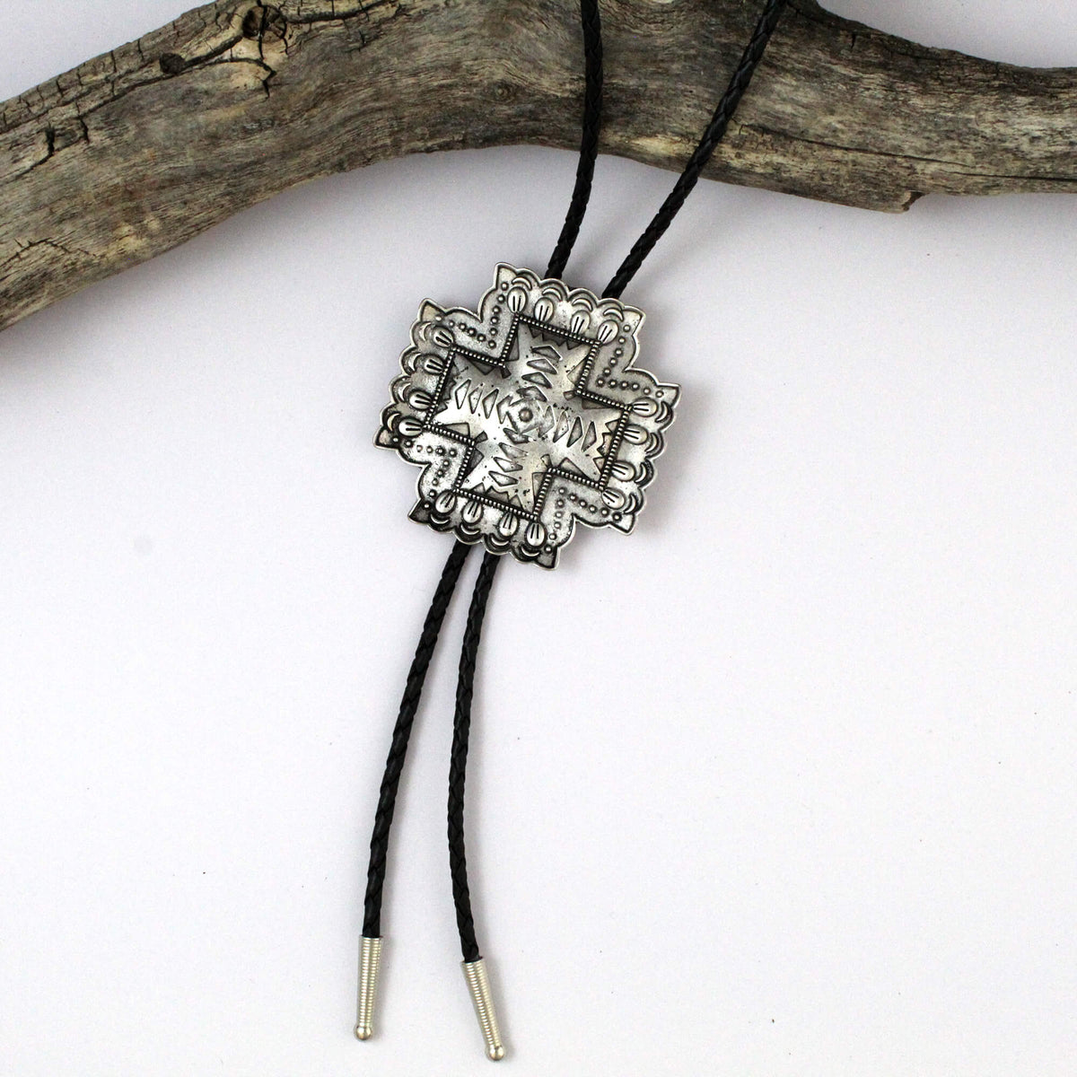 A silver-toned cross bolo tie pendant with engraved details, suspended on black cords with silver-toned tips, displayed against a white background with a piece of driftwood at the top.