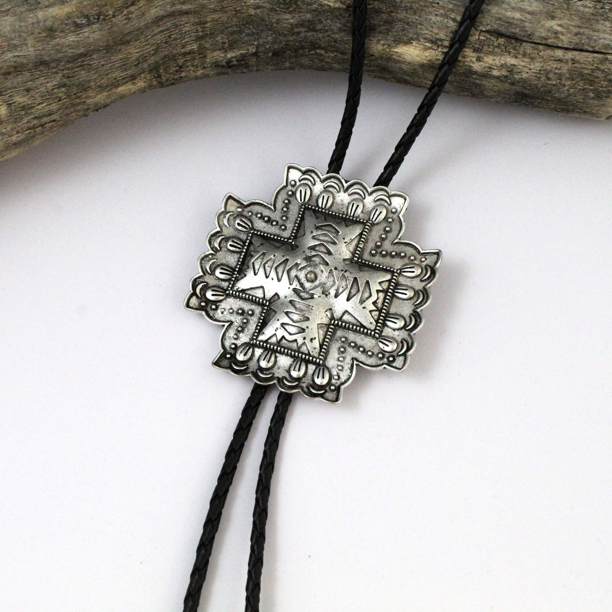 A silver-toned western-style bolo tiefer pendant necklace with an engraved star and cross design, suspended on a black cord against a white background with a piece of driftwood at the top.