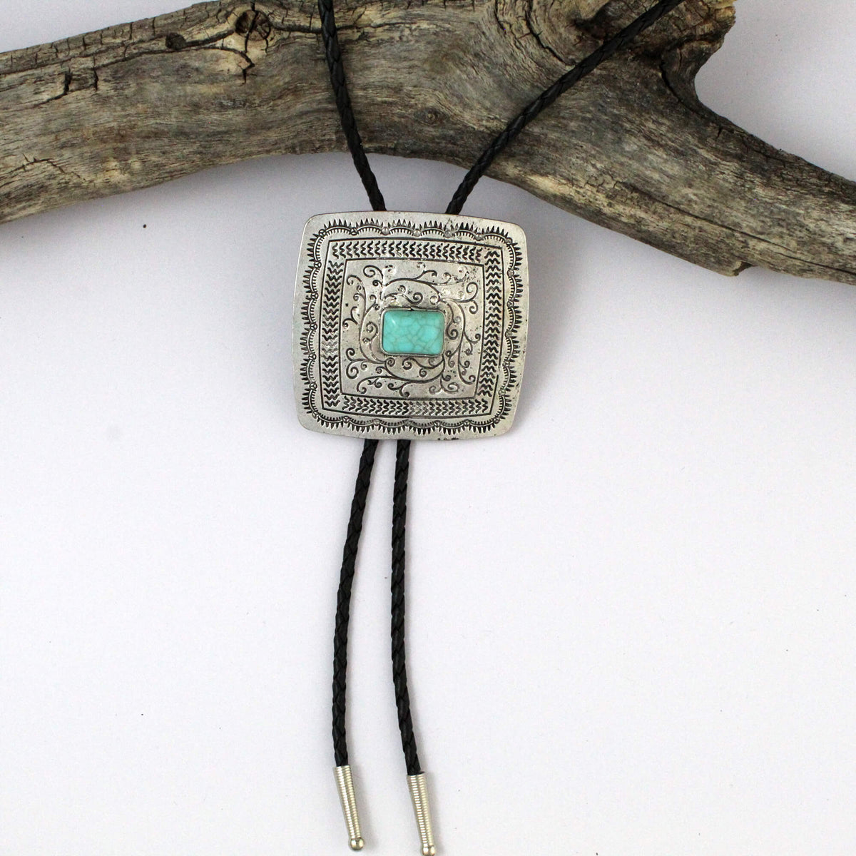 A square silver-toned bolo tie pendant necklace with a turquoise-colored center stone, hanging from black braided cords with silver-toned tips, displayed against a white background with a piece of driftwood.
