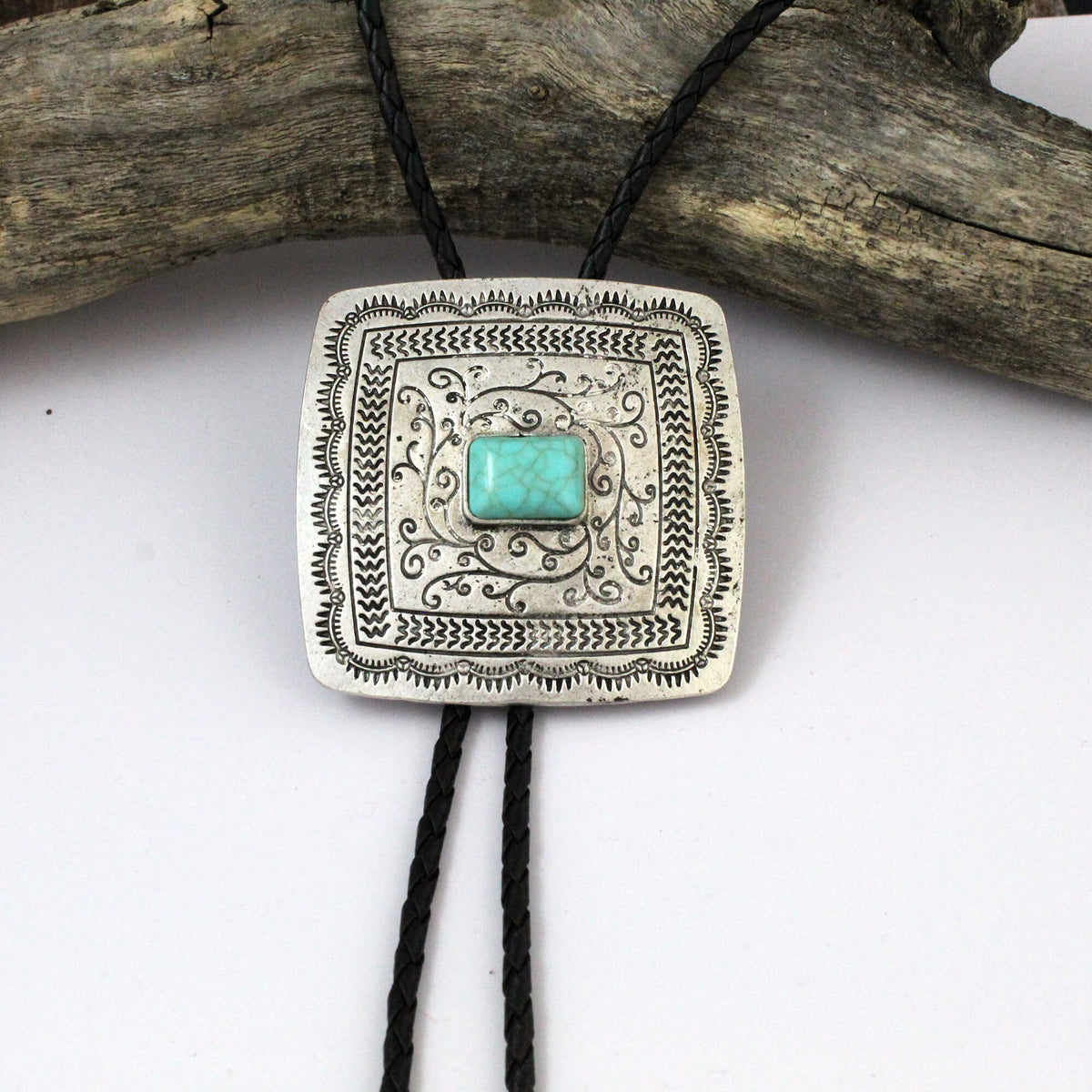 A square silver bolo tie necklace with an engraved pattern, featuring a central turquoise stone and hanging from a black cord, displayed against a white background with a piece of driftwood.