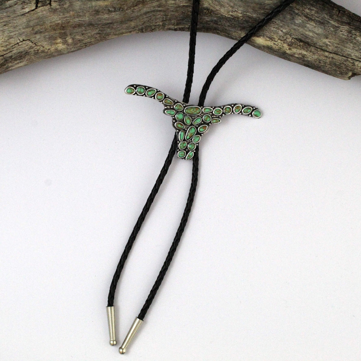 A silver-toned bolo tie necklace featuring a longhorn-shaped centerpiece paved with small green stones, suspended on a black braided cord with silver-toned metal tips, displayed on a white surface with a piece of driftwood at the top.