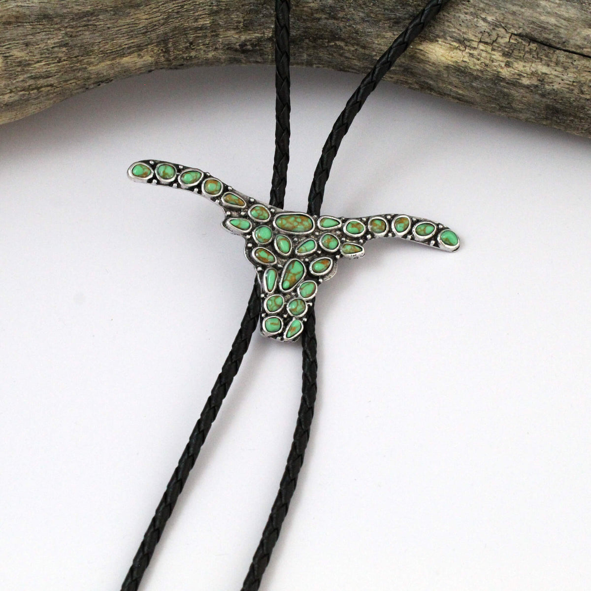 A silver-toned longhorn pendant necklace embellished with small green stones, hanging from a black braided cord on a white background with a piece of driftwood at the top.