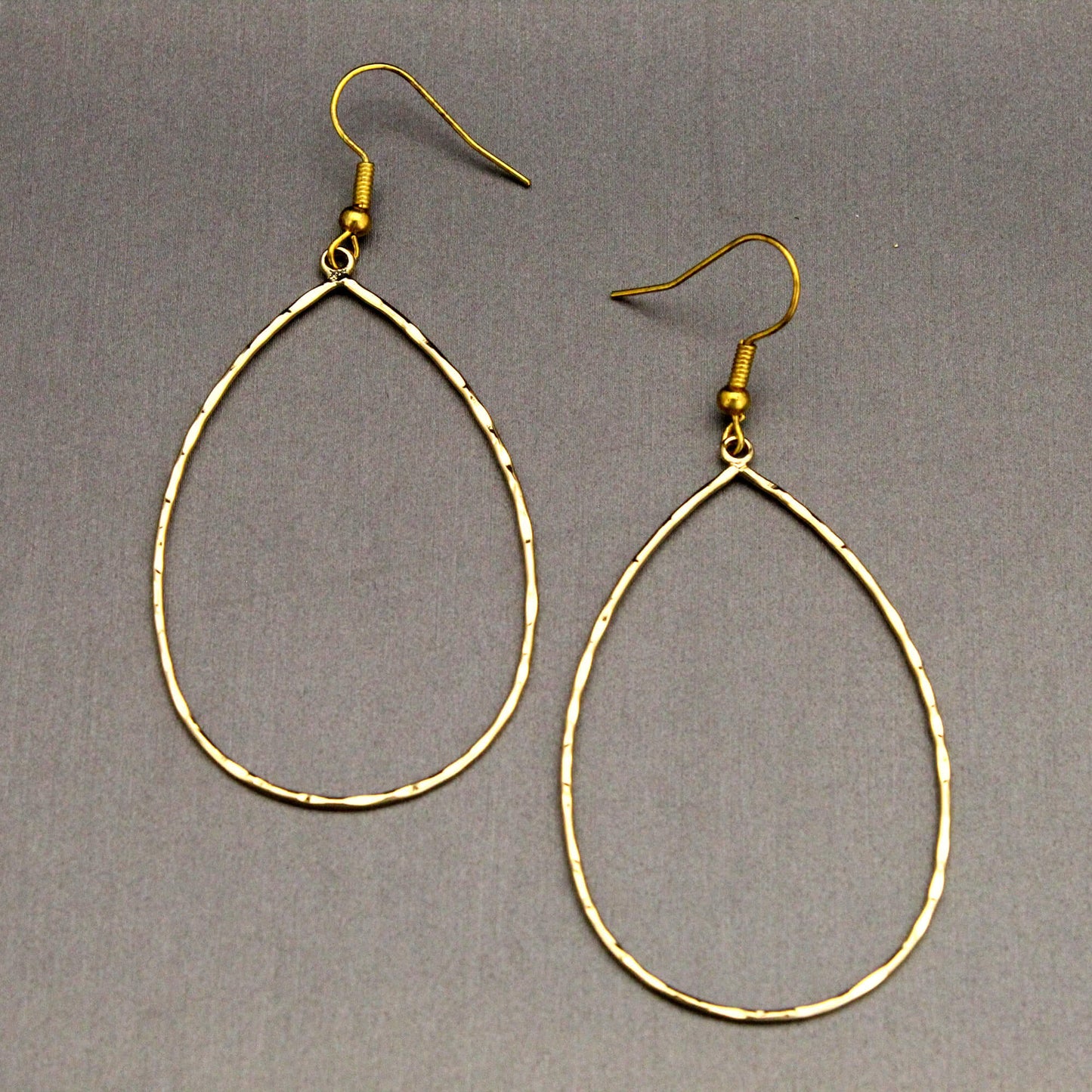 A pair of gold-tone teardrop hoop dangle earrings with textured, hammered edges and fishhook backings, displayed on a neutral gray background.