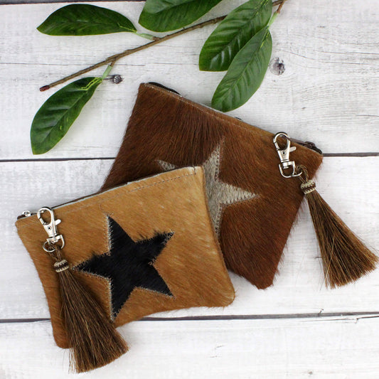 Two small cowhide pouches with tassel zipper pulls and silver clasp hardware, arranged on a white wood surface with green leaves in the corner. One pouch is tan with a black star motif, and the other is medium brown with a beige star motif.