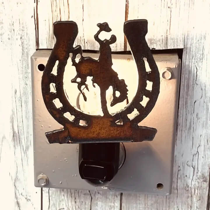 A rustic metal horseshoe-shaped night light featuring a cowboy silhouette, mounted on a square metal backplate with a black outlet insert, set against a whitewashed wood background.