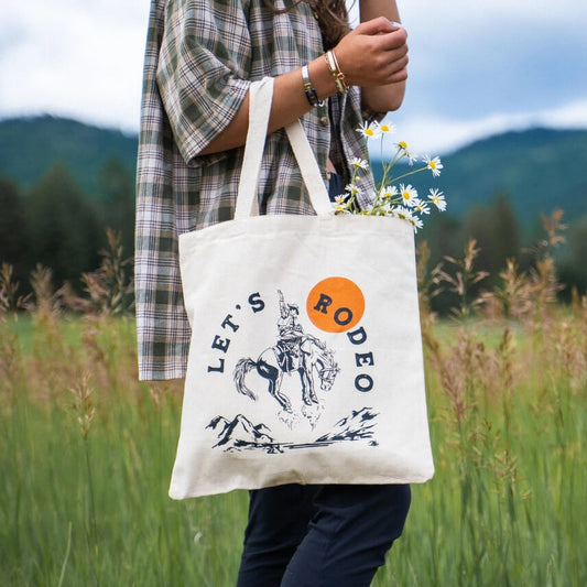 Person holding a tote bag with 'Let's Rodeo' design in a natural setting