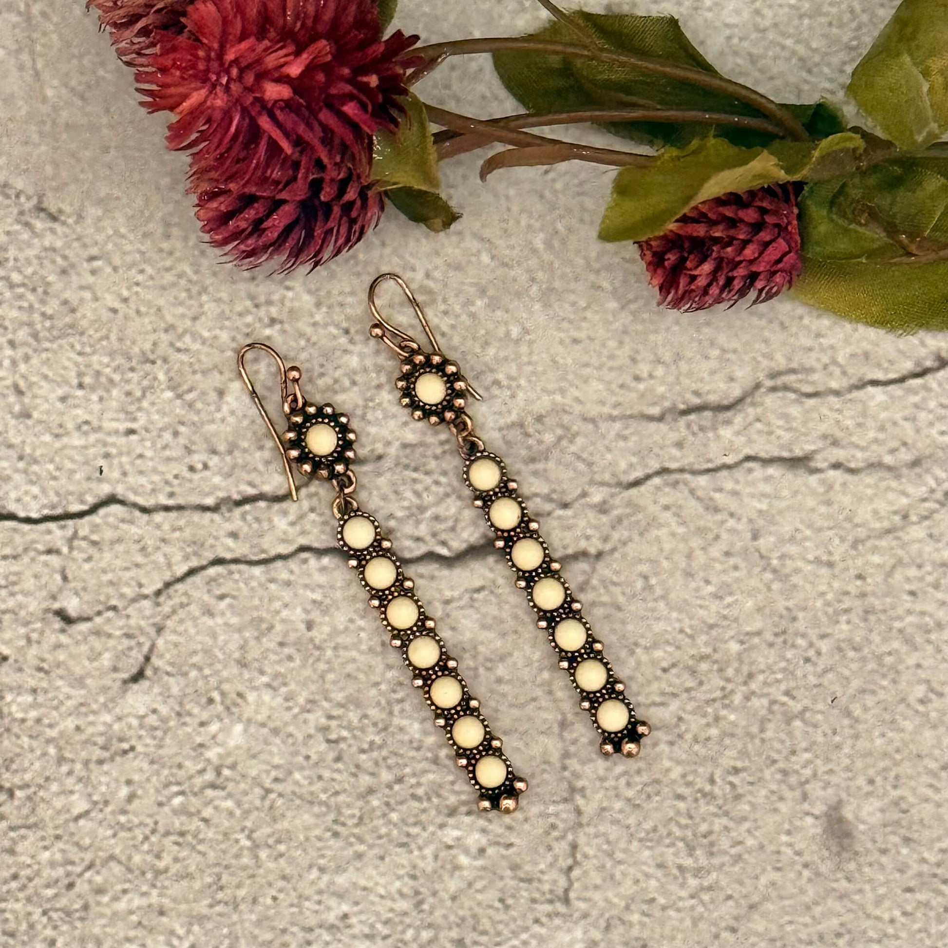 Pair of long earrings with circular beads on a textured surface with flowers in the background