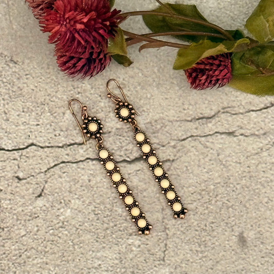 Pair of long earrings with circular beads on a textured surface with flowers in the background