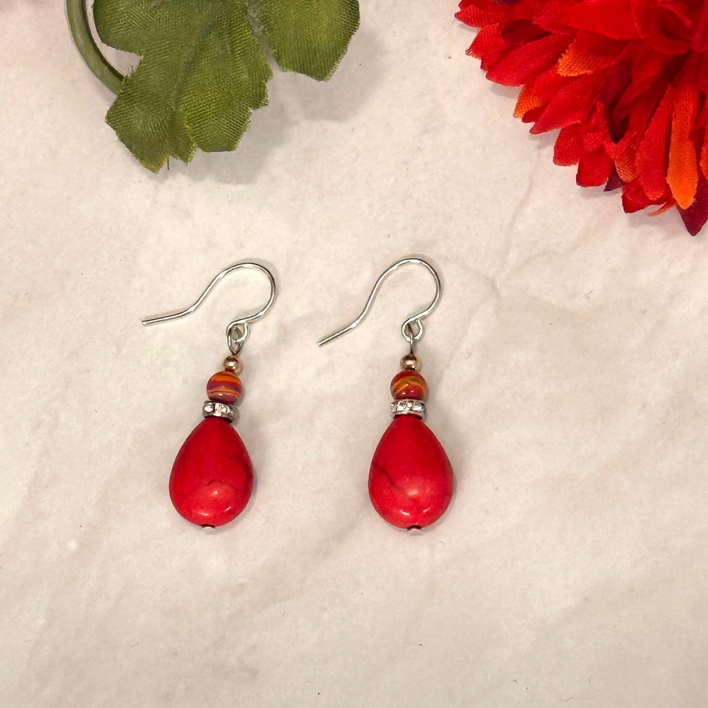 Red teardrop earrings with silver hooks on a textured surface with a red flower and green leaf in the background.