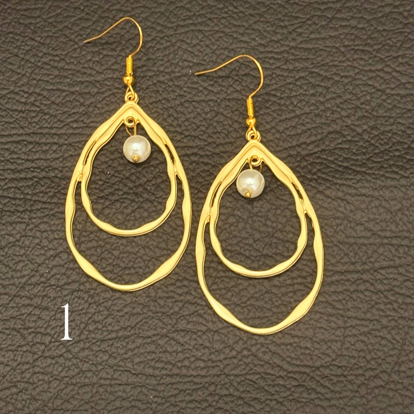 A pair of gold-toned dangle earrings with irregular double teardrop-shaped rings and a small white pearl at the center, displayed on a dark textured leather background.