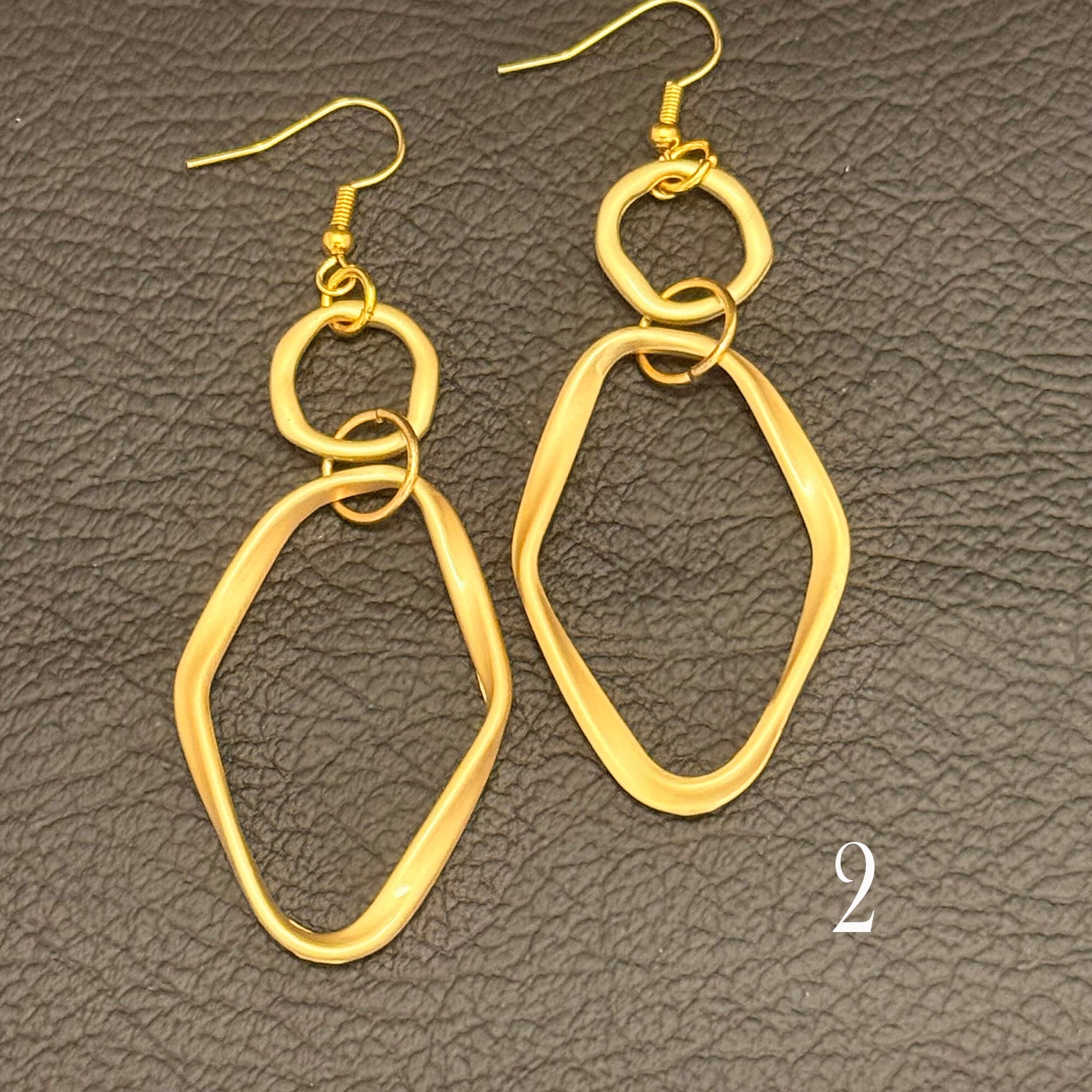 A pair of gold-toned dangle earrings with irregular open geometric shapes and fishhook wires, displayed on a textured dark gray surface. A small white number '2' appears in the lower right corner.