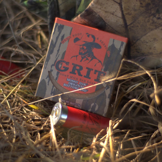 A box of Grit Goat Milk Bar Soap on dry grass with a single red and silver shell placed in front of it.