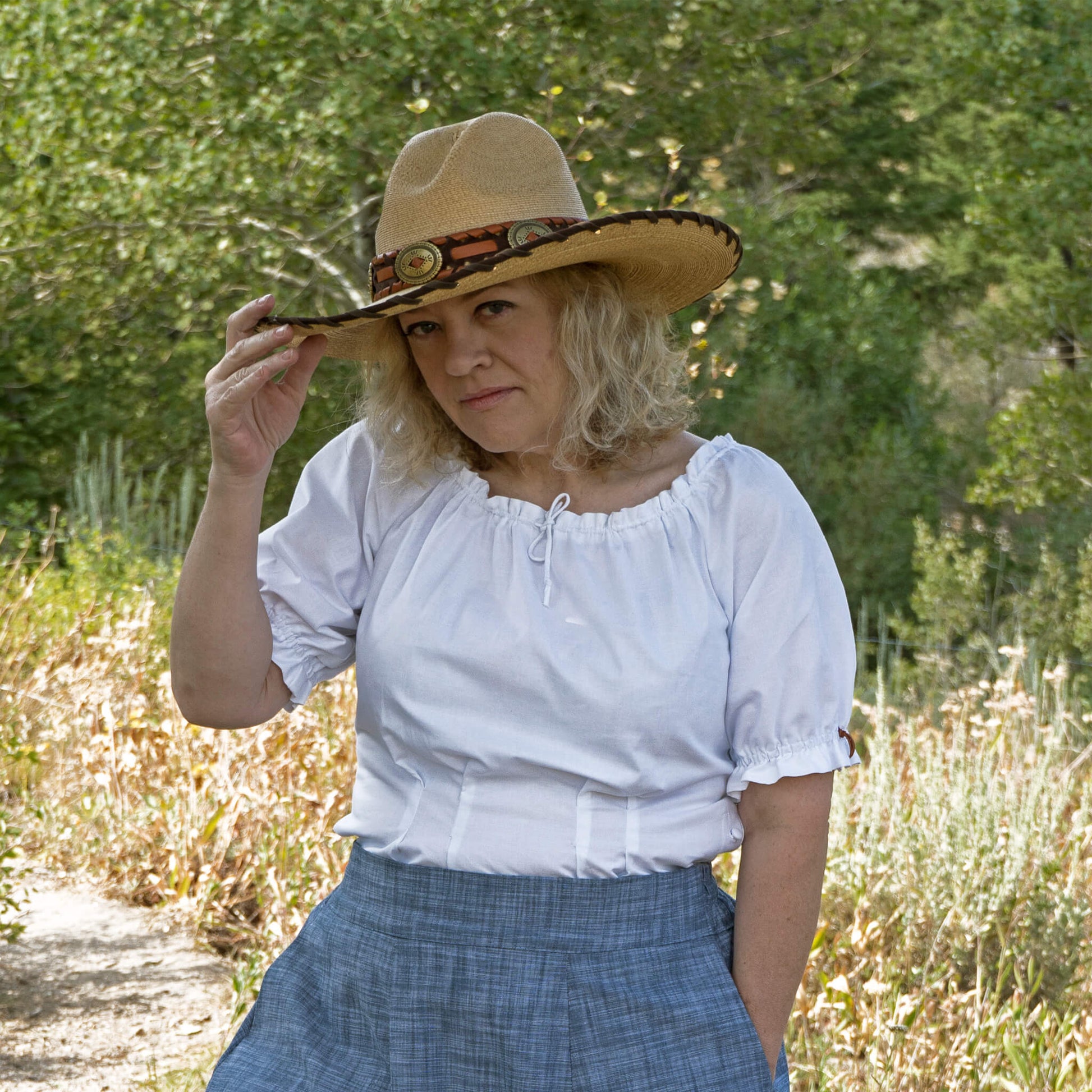 Looking for a fun flirty summer blouse to wear with your hat and boots? Then the Olmstead Blouse is the perfect look for you. Blouse has a drawstring neckline so you have control over how open a neckline you are feeling. Lower left side opening with covered button closure. Elbow length sleeves have an elastic fit with removable leather tie. Wear it tucked in or out. Wear it off the shoulder or dress it up with a stunning necklace. You can alter the look to your desire. 100% Cotton. Made in USA