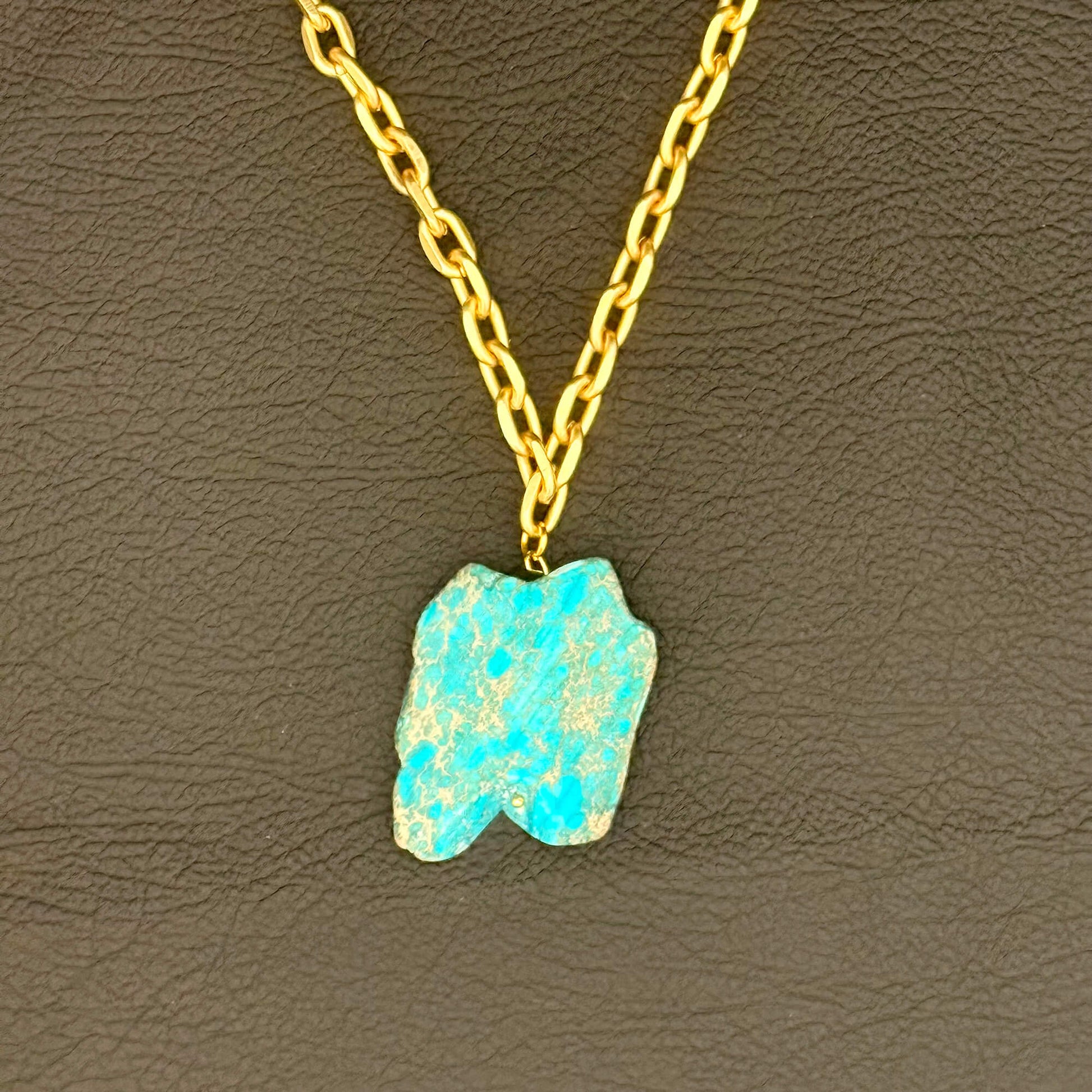 A gold chain necklace with a raw, irregular-shaped turquoise-blue stone pendant, displayed on a textured dark brown leather background.