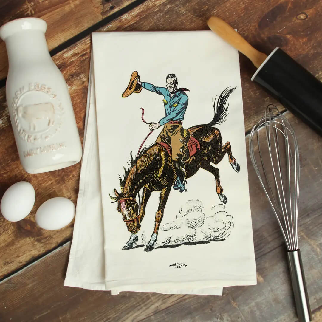 A folded white kitchen towel with a vintage-style illustration of a cowboy riding a bucking horse, styled on a rustic wooden table with a white milk bottle, eggs, a rolling pin, and a whisk.