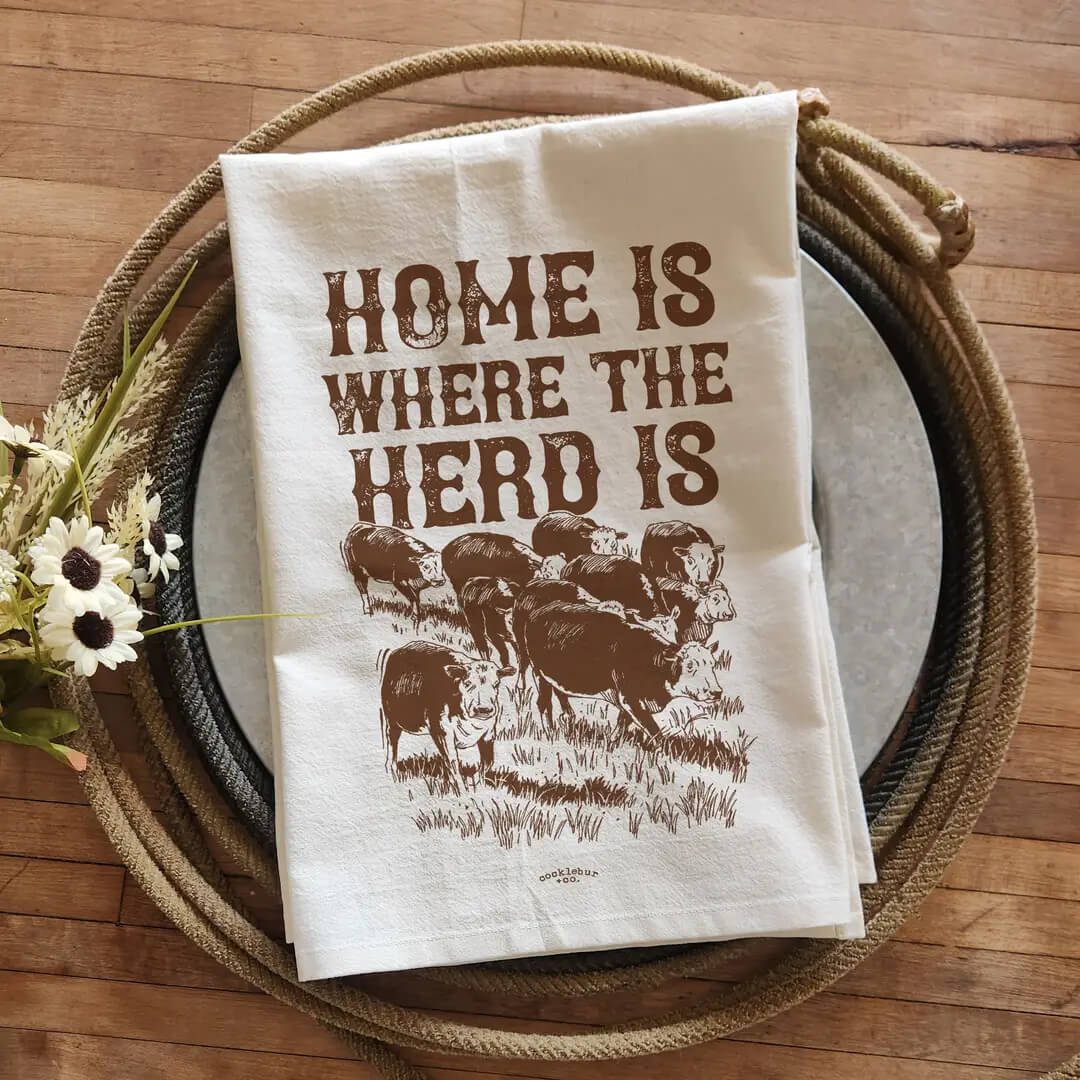 An off-white cotton tea towel with a brown graphic print reading 'HOME IS WHERE THE HERD IS' and showing a herd of bison, styled on a rustic wooden table with a woven placemat and white plates.