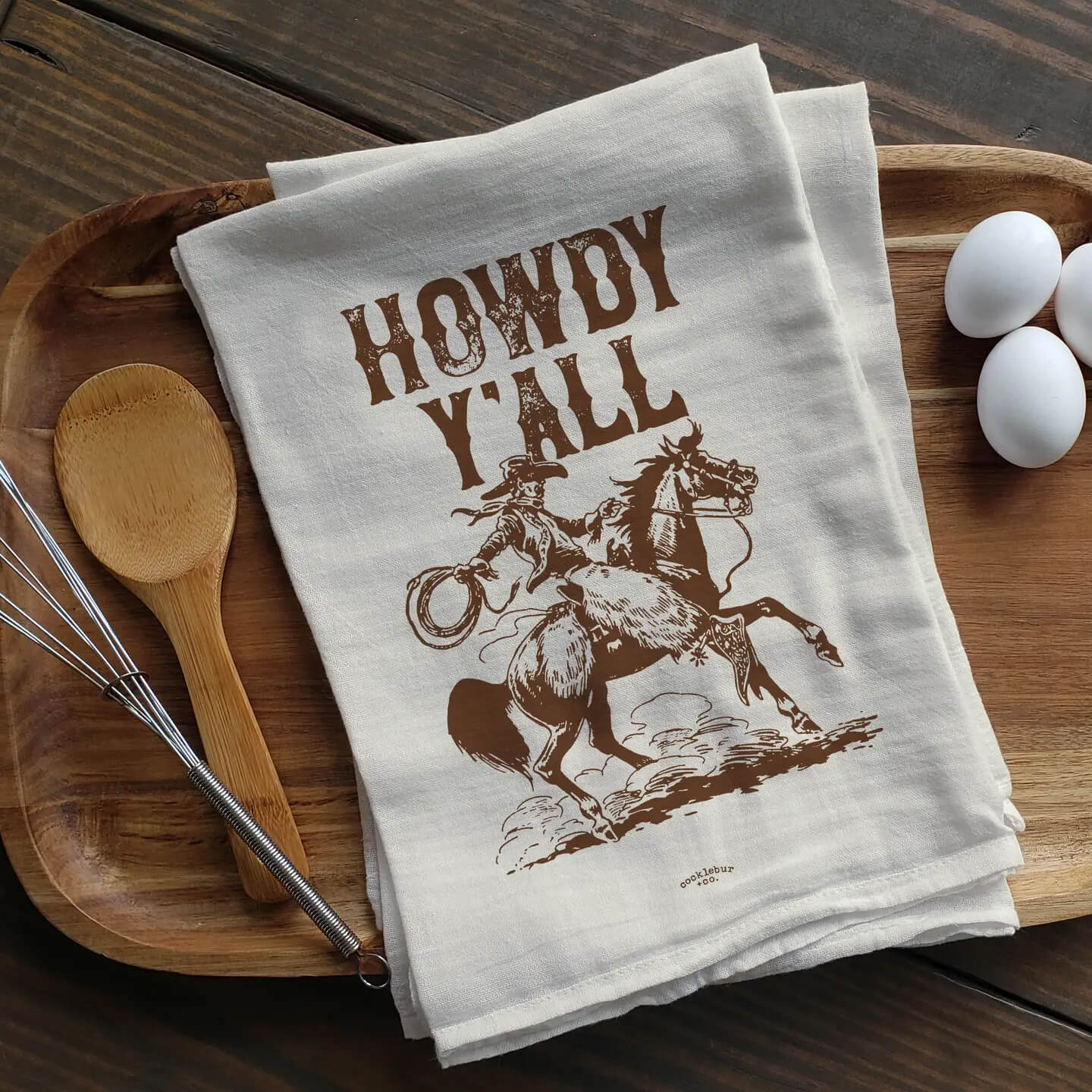 A folded cream-colored kitchen towel with a brown graphic of a cowboy on a bucking horse and the words 'HOWDY YALL' printed on it, styled on a wooden tray with a wooden spoon, metal whisk, and white eggs on a dark wood table.