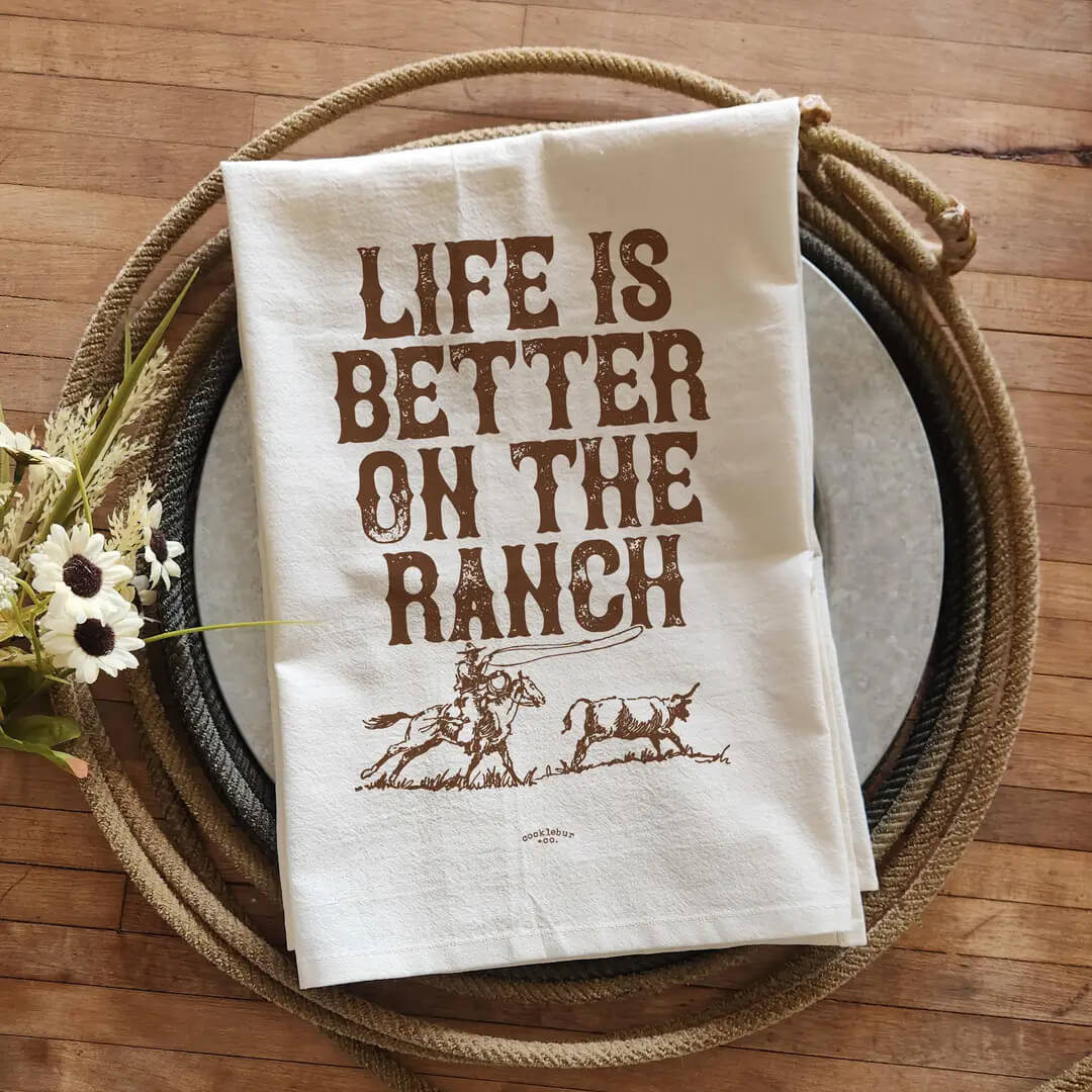 An off-white cotton tea towel with brown printed text reading 'LIFE IS BETTER ON THE RANCH' and a cowboy riding a horse beside a steer, displayed on a rustic wooden table with a coiled rope and metal platter.