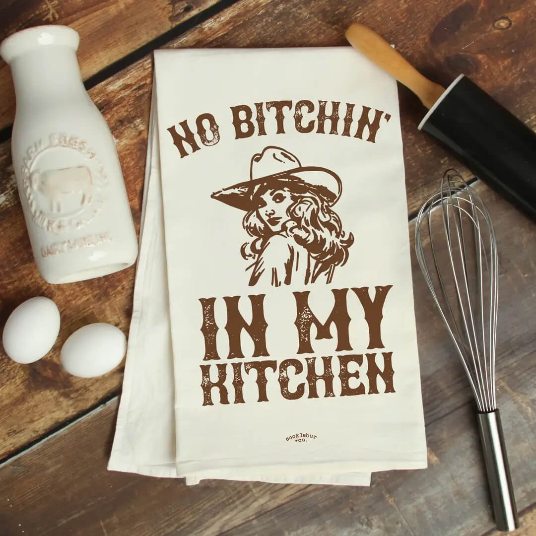 A white kitchen tea towel with a brown graphic print reading 'NO BITCHIN' IN MY KITCHEN' and showing a woman in a cowboy hat, laid flat on a rustic wooden table with baking props including a milk bottle, eggs, rolling pin, and whisk.