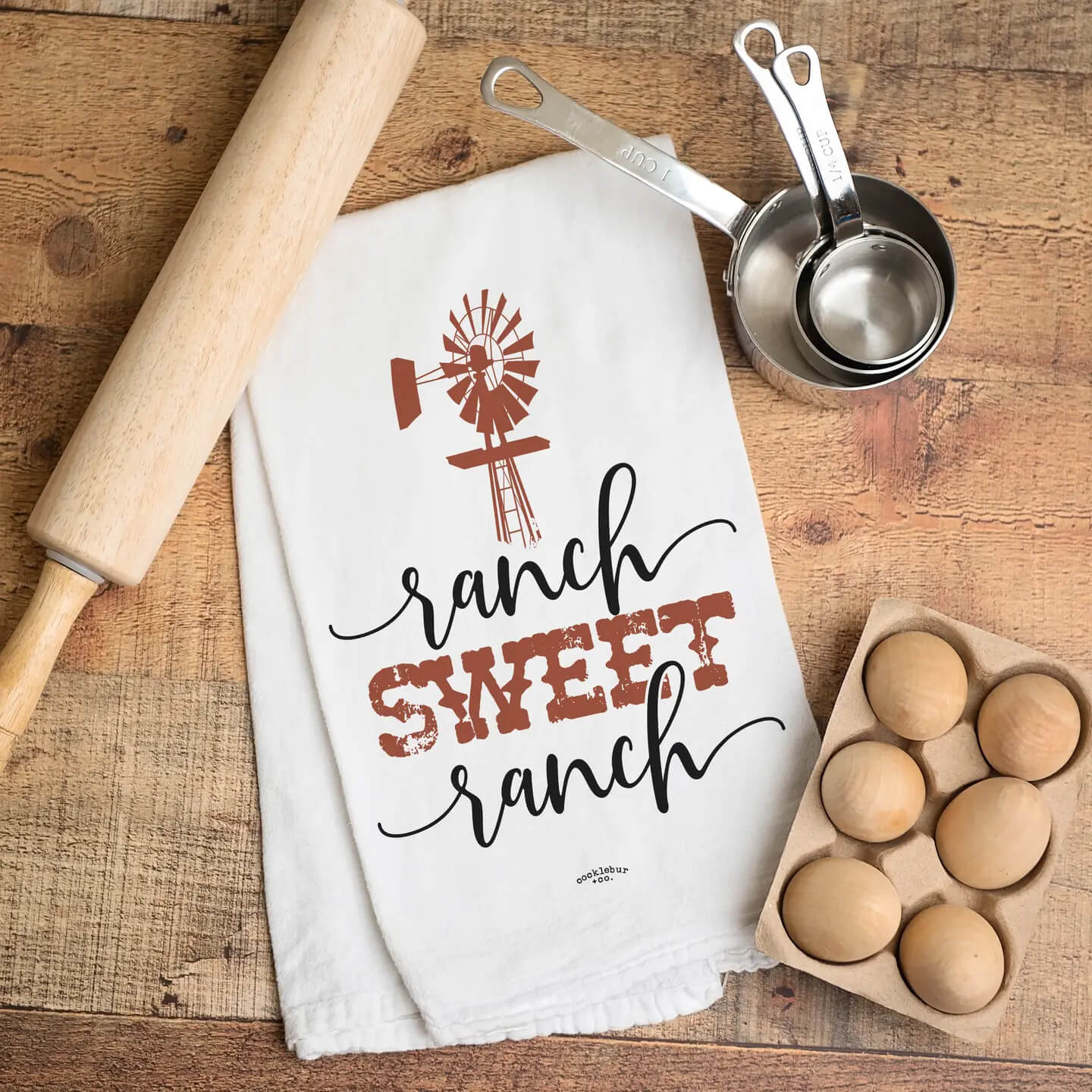 A white kitchen towel with a rustic 'ranch sweet ranch' design in black and terracotta red, laid flat on a wooden table alongside a wooden rolling pin, metal measuring cups, and a carton of eggs.