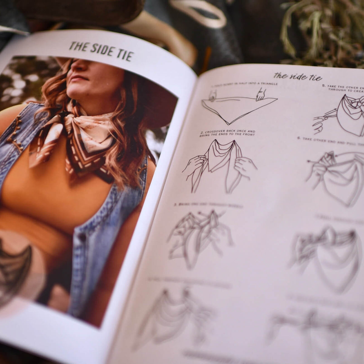 An open book showing a fashion guide page titled 'THE SIDE TIE' with a photo of a woman wearing a side tie and black line drawings of how to tie it. The book is photographed in a styled setting with warm lighting.