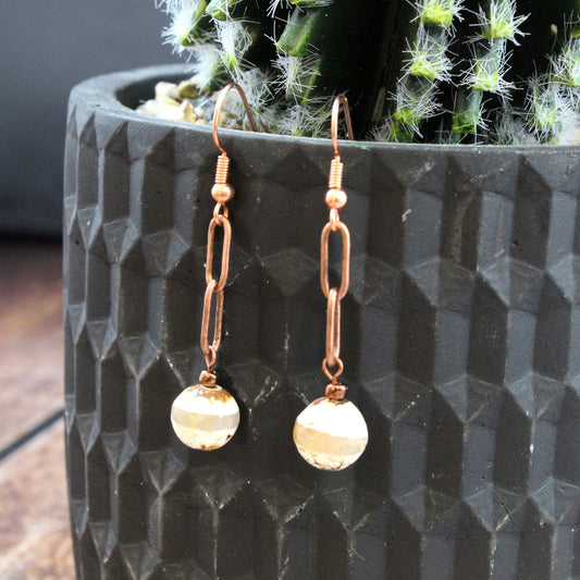 A pair of copper drop earrings with faceted clear beads, hanging in front of a textured black planter containing a cactus.