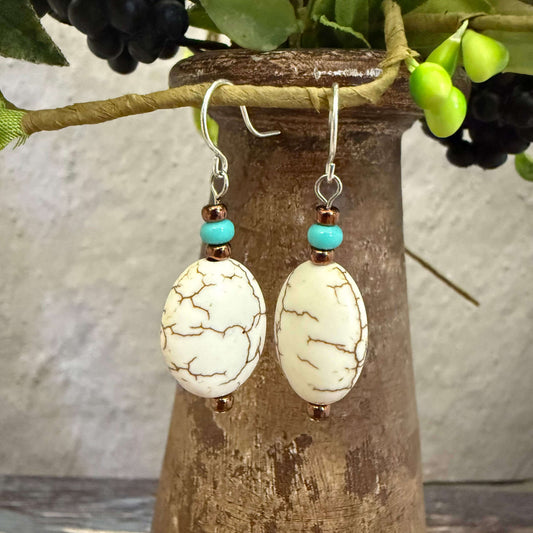 Pair of earrings with white beads and turquoise accents hanging on a wooden stand.