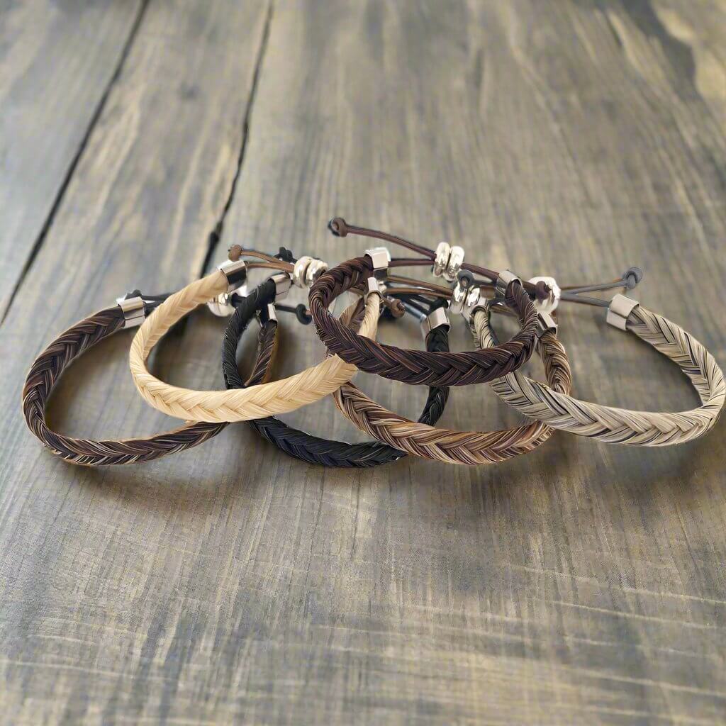 Five braided horsehair bracelets in various shades of brown, beige, and black, arranged on a rustic wooden surface.