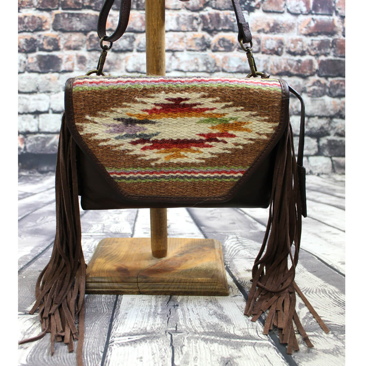 Ariat Sheridan crossbody bag is genuine leather and God’s eye southwestern pattern on handwoven fabric on the front flap. Brown suede fringe on the sides.&nbsp;Snap closure with one zipper pocket inside.&nbsp;Measures: 9” X 7” X 1”. Imported.