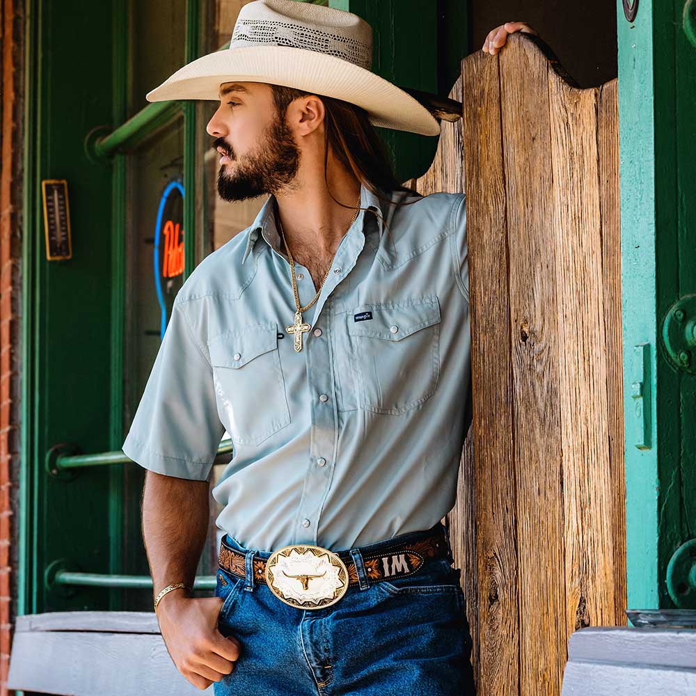 Man wearing a cowboy hat and western attire leaning against a wooden post.