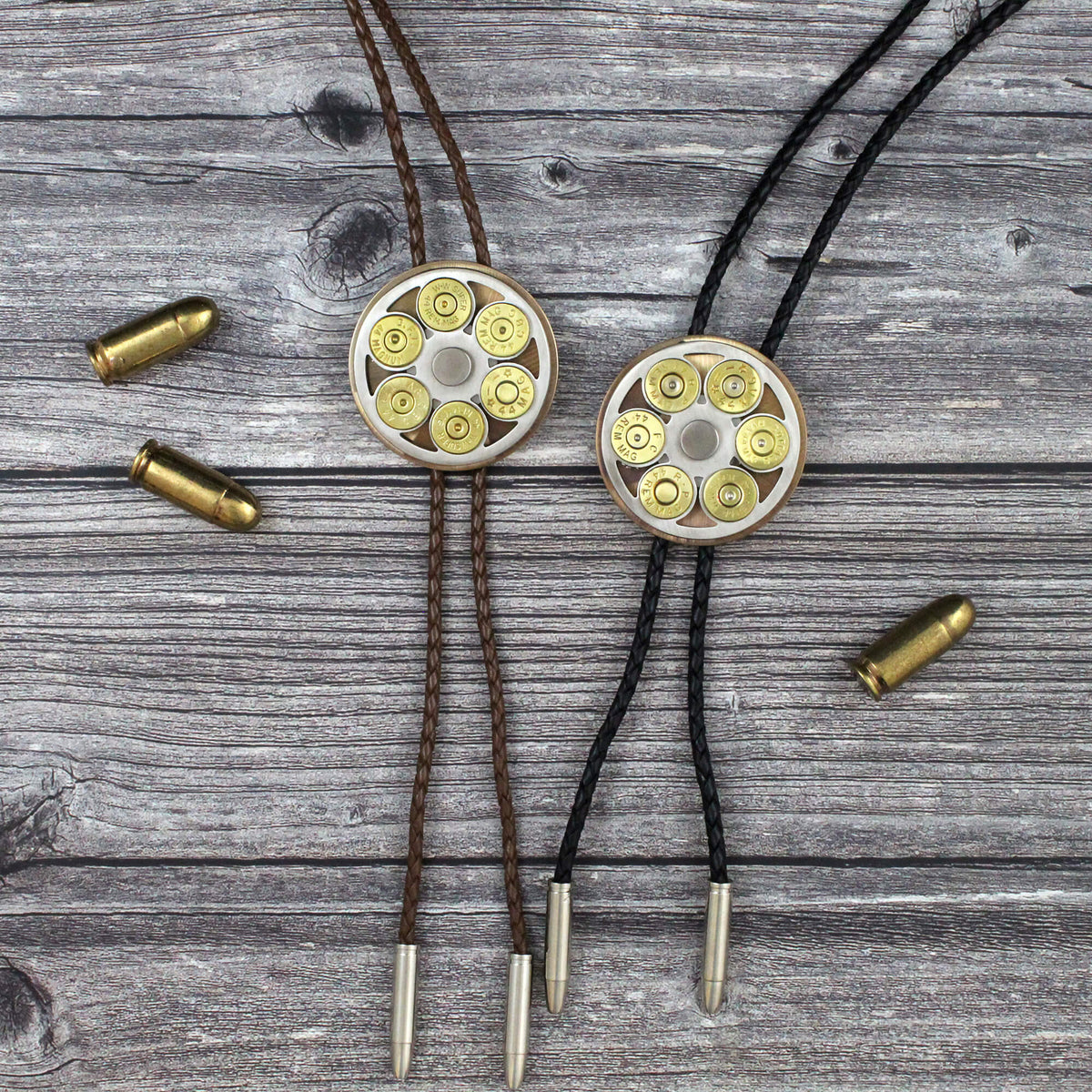 Two shotgun shell bolo ties on a weathered gray wood background, one with brown cords and one with black cords, each featuring a circular metal cylinder filled with brass and silver bullets, with loose bullets scattered nearby.