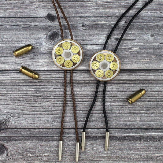 Two shotgun shell bolo ties on a weathered gray wood background, one with brown cords and one with black cords, each featuring a circular metal cylinder filled with brass and silver bullets, with loose bullets scattered nearby.