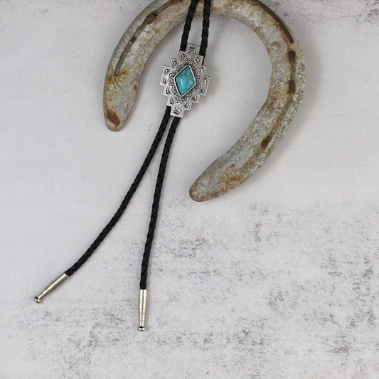 A silver-toned bolo tie with a turquoise stone centerpiece and black braided cords, displayed on a rustic white surface with a weathered horseshoe prop.