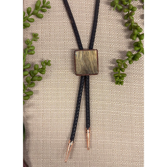 Bolo tie with square wooden pendant on a beige fabric background with green leaves