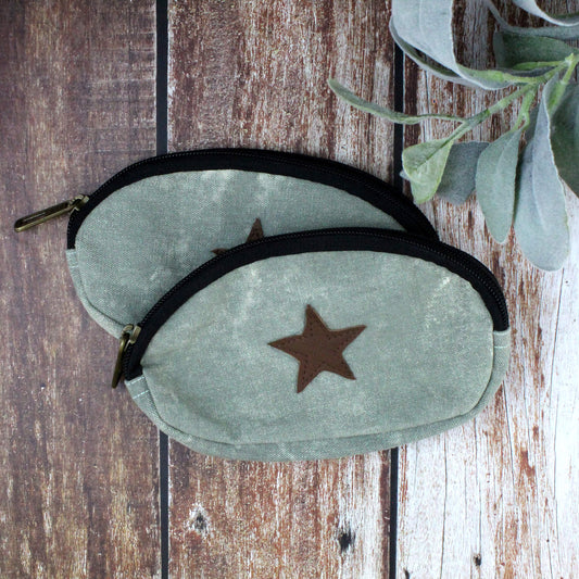 Two sage green canvas zippered pouches with brown star patches and black zippers, arranged on a rustic wooden surface with green leaves in the corner.