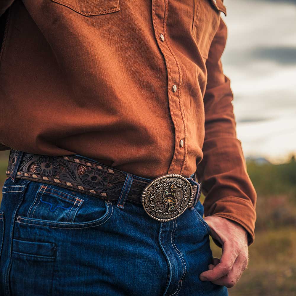Person wearing a brown shirt and blue jeans with a decorative belt buckle outdoors.
